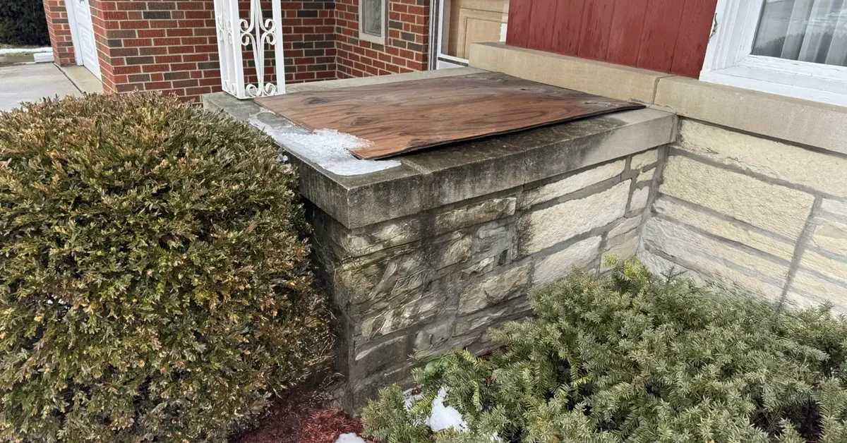Damaged brick wingwall with weathered mortar joints and a boarded-up cap before masonry repair